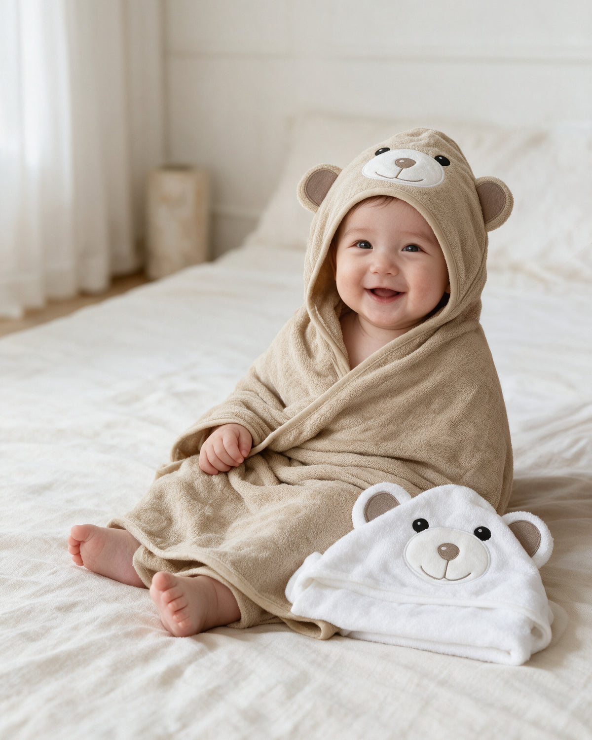 Baby sitting on a bed in the Cream Bear hooded towel, with the Little Bear towel beside